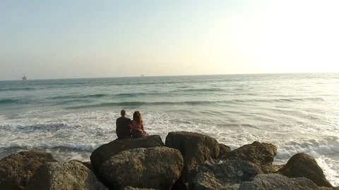 Couple Looking at Ocean While Sitting On Rocks Aerial 4k Stock Footage 79665070
