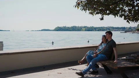Couple in love sits on bench under tree at promenade, admires view of Lake Garda 스톡 동영상 127542017