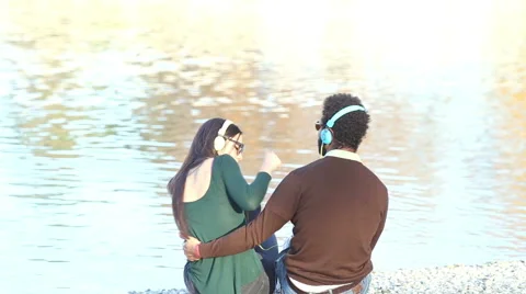 Couple in love sitting by lake and listening to music with headphones Stock Footage 59555898