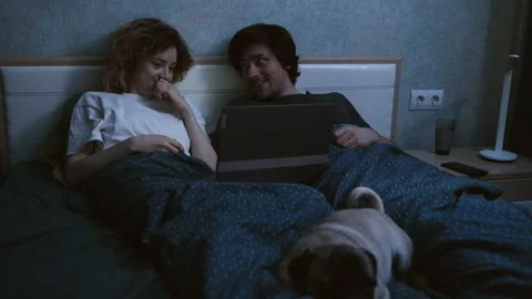 A couple is lying in bed and looking in a laptop, discussing and laughing. Stockbeeldmateriaal 197558156