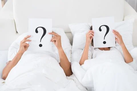Couple Lying On Bed Holding Question Mark Over Faces Stock Photos