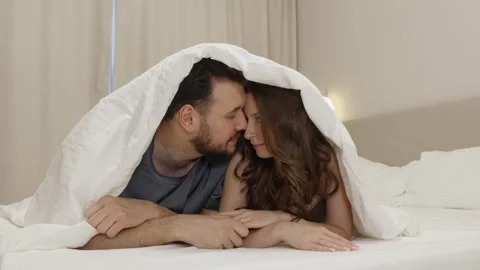 Couple lying in bed under blanket, holding hands and kissing Stock Footage 310766162
