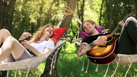 Couple lying on hammocks, talking and laughing. Young man with guitar, woman Video stock 158923754