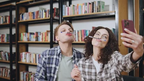 Couple playfully posing with pens in a library while taking a selfie amid Stock Footage 308667159