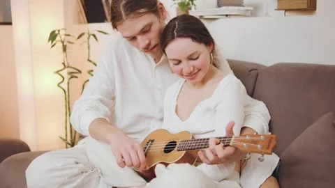 Couple plays ukulele on sofa while a cat sits nearby in a cozy room during Stock Footage 322805813