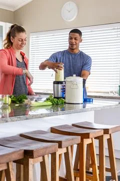 Couple preparing green drink at kitchen island, man in blue shirt, woman coral Stock Photos