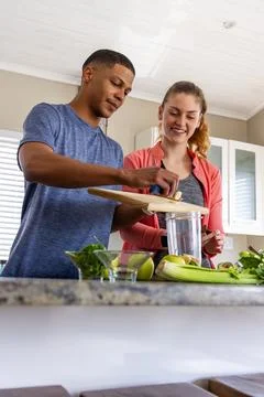 Couple preparing smoothie in kitchen, man holding cutting board while woman Stock Photos