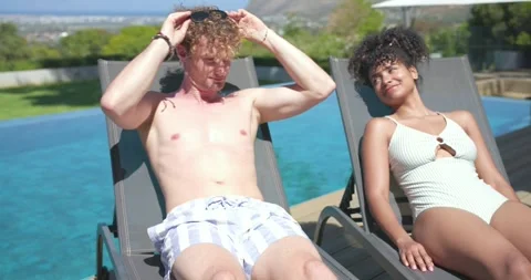 Couple reclining on pool deck, man removing shades, sparking glance, holding Stock Footage 344333914