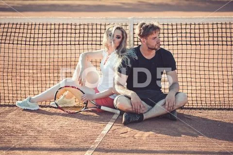 Couple relax on tennis court after training. Sportsman and sportswoman sit at Stock Photos