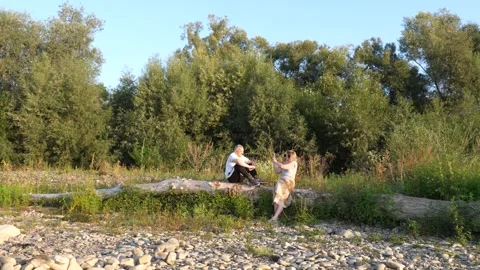 A couple relaxes by the river, sitting on a fallen tree and taking photos aga Stock Footage 341864132