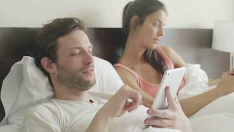 Couple relaxing in bed chatting while using personal wireless devices Stock Footage 75177344
