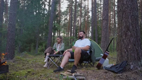Couple relaxing by a campfire in a pine forest campsite Stock Footage 327607481
