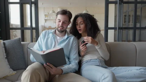 Couple Relaxing On Sofa, Reading Book Together, Reacting With Surprise Interest Stock Footage 303759753