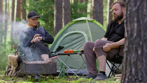Couple relaxing while grilling meat at a forest campsite Stock Footage 323606739