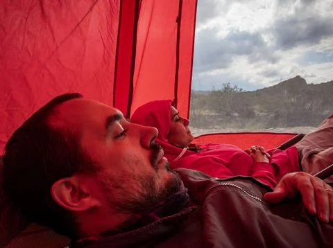 Couple rests on deck chairs inside a large red tent in a mountain environment Stock Photos