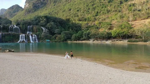 Couple Run Down the Beach with Stunning Ban Gioc Waterfall in Vietnam Behind Stock Footage 125497679