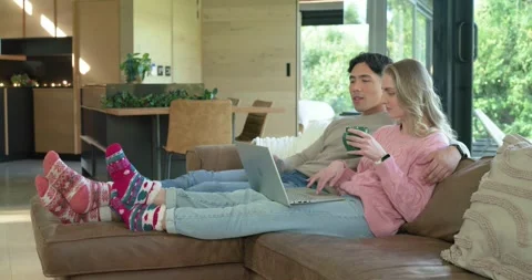 Couple settling on sectional sofa, typing on laptop, sipping mugs, chatting in Stock Footage 341931931