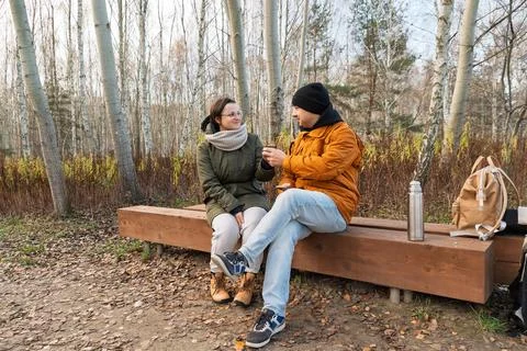 Couple sharing drink while sitting on bench in birch grove Stock Photos