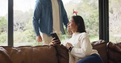 Couple sharing phone on sofa as man entering donning Santa hat prompts woman in Stock Footage 341467433