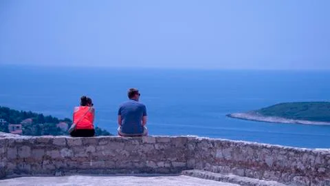 Couple sit high up on a wall looking out at the ocean view Stock Photos