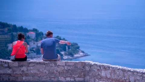 Couple sit high up on a wall looking out at the ocean view Stock Photos