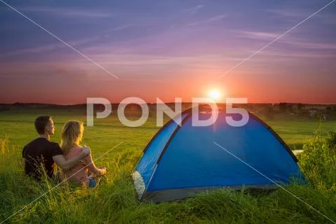 The couple sit near camping tent by sunset (sunrise) background. Wide angle Stock Photos