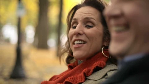 A couple sit at a park bench and talk as the woman laughs on a nice fall day Stock Footage 98380812