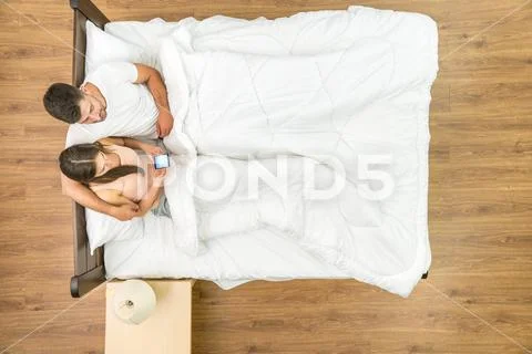 The couple sit with a phone on the bed. View from above Stock Photos