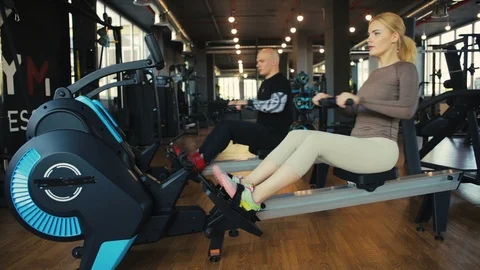 Couple sit on rowing machines and pull oars synchronously Stock Footage 110575387