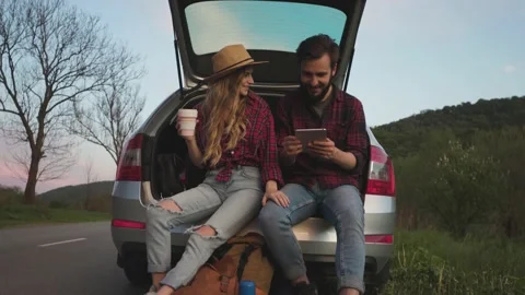 Couple sit on the trunk of car and wonder at photo on tablet Stock Footage 131458996