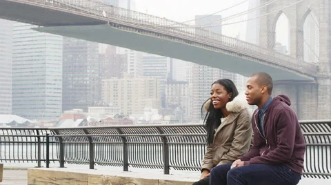 A couple sits on a bench in a park in front of the Brooklyn bridge on an Stock-Footage 45762033