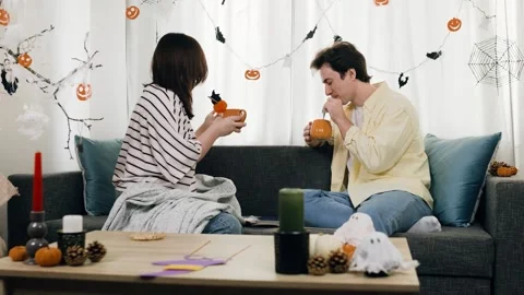 Couple sits on sofa drinking pumpkin mugs for Halloween celebration. The man and Stock Footage 346829905