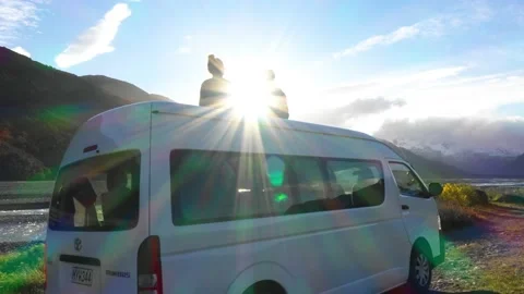Couple sits on Van in Mountain Range setting as Sun sets between them - Aerial Stock Footage 130358290