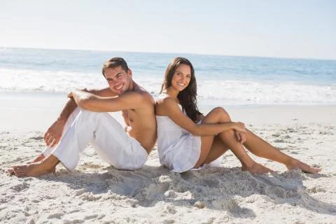 Couple sitting back to back on the sand smiling at camera Stock Photos