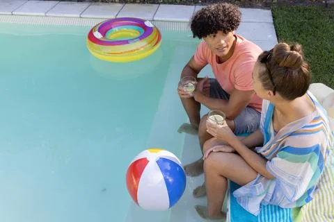 Couple sitting at backyard pool edge, holding drinks, pool ring, beach ball Stock Photos