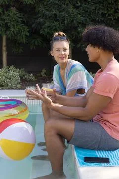 Couple sitting at backyard pool edge, feet in water, holding drinks, colorful Foto stock