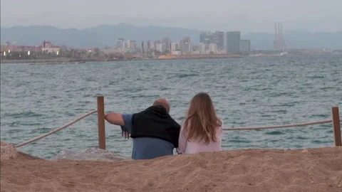 Couple sitting on beach in front of the sea with a panoramic view of city Stock Footage