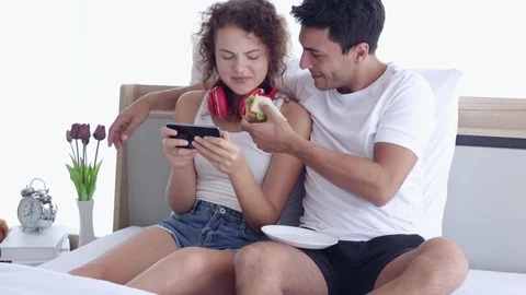 Couple sitting on the bed. A man eating the sandwich at breakfast. A woman liste Видео 107735520