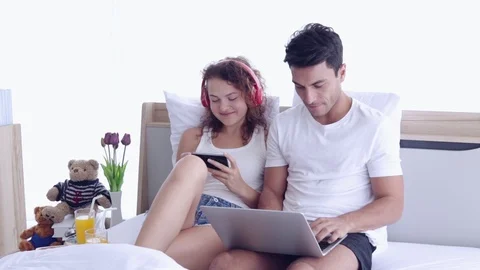 Couple sitting on the bed. Man typing laptop and woman listening to music by sma Stock Footage 107735931