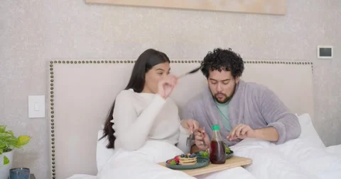 Couple sitting on bed sharing breakfast tray, woman waving fork offering bite, Stock Footage 345589149
