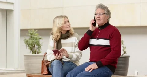 Couple sitting on bench in courtyard hearing smartphone ringtone, man answering Stock Footage 342492502