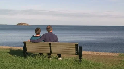 A couple sitting on a bench looking out to a large rock in the ocean (1 of 2) Stock Footage 10775144