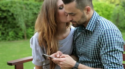 Couple sitting on the bench. Man is using phone and woman is kissing him Stock Footage 51440965