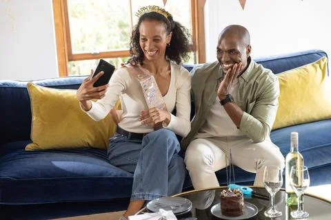 Couple sitting on blue sofa taking selfie with smartphone, wearing birthday sash Stock Photos