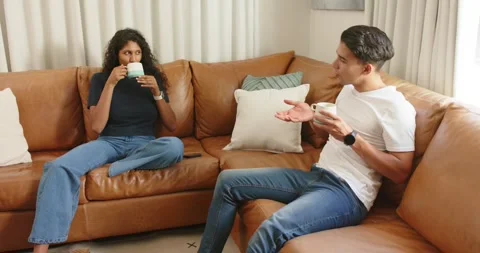 Couple sitting on brown sofa, man starting relaxed chat while woman lowering Stock Footage 342019194