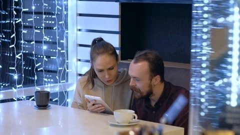 A couple sitting in a cafe, talking, drinks coffee and watching in smartphone. Video stock 121869967