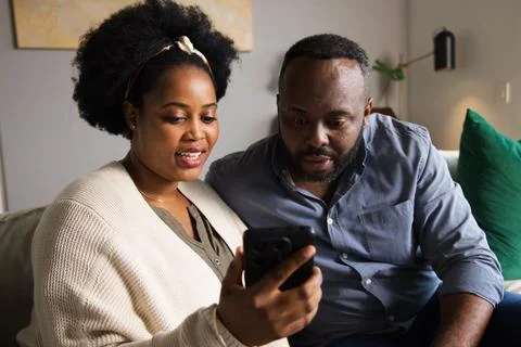 Couple sitting closely on light-colored sofa sharing black smartphone, woman Stock Photos