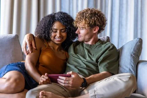 Couple sitting closely on light-gray sofa in living room, man holding pink Fotos Stock