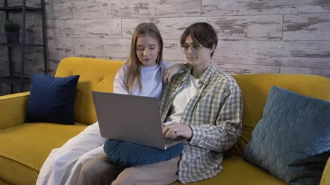 Couple is sitting on the couch, looking at a laptop and talking, a man is typing Video stock 259793618