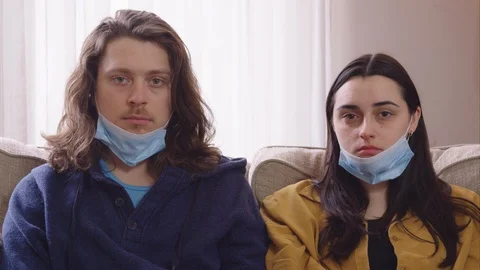 Couple sitting on a couch put on N95 masks and look into camera emotionless - Stock Footage 129468005
